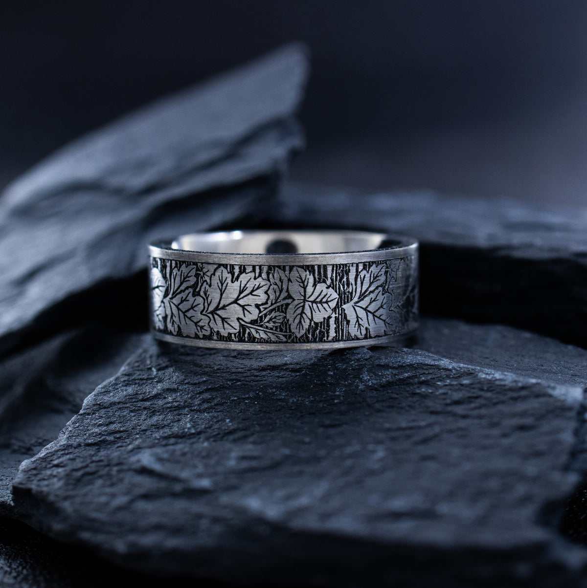 A silver band ring with an engraved leaf pattern, resting on dark slate stones against a dark, moody background.
